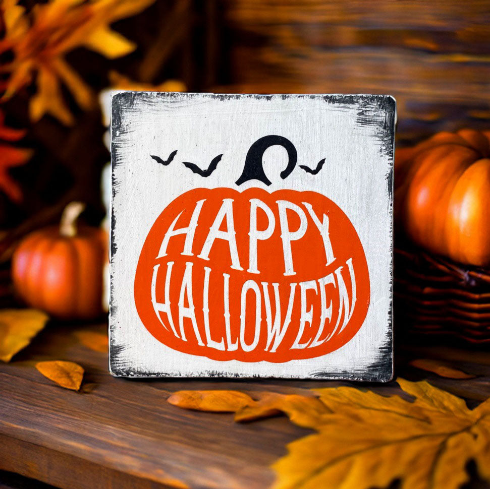 Kleines weißes Holzschild mit einem orangen Kürbis, der den Schriftzig Happy Halloween enthält. Um den Kürbis sind 3 kleine schwarze Fledermäuse.